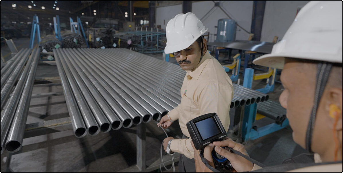 A Meta Engitech inspector in a white hard hat taking a handheld measurement from a rack of finished precision steel tubes on the shop floor, a colleague working alongside him.