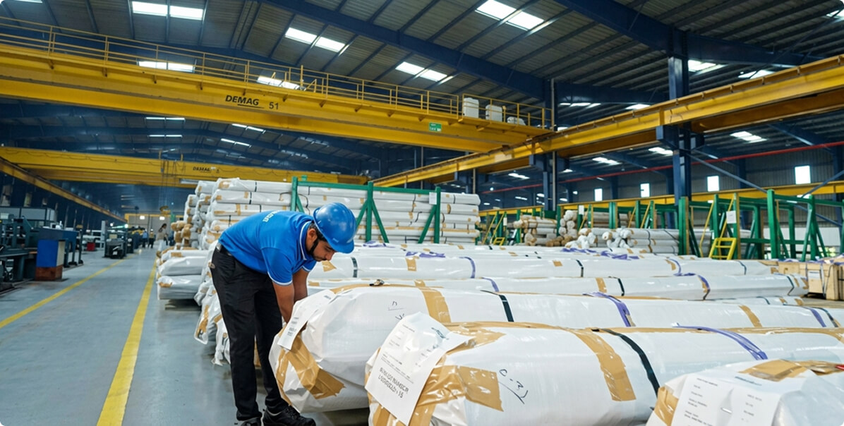 A Meta Engitech operator in a blue hard hat checking wrapped and strapped bundles of finished tube in a wide despatch bay, overhead gantry cranes running the length of the roof.